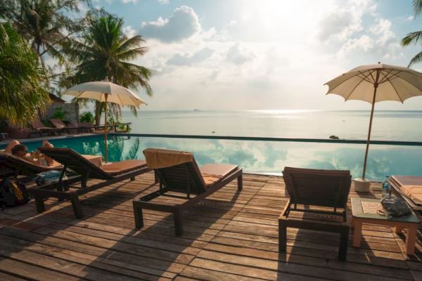 A serene poolside with lounge chairs, umbrellas, palm trees, and a view of the ocean under a partly cloudy sky.