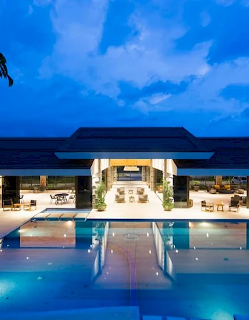 A luxurious building with large open spaces is illuminated at night, featuring a pool reflecting the structure and the evening sky.