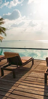 Poolside scene with lounge chairs, parasols, and a serene view of palm trees and the ocean reflecting the sky on a sunny day.