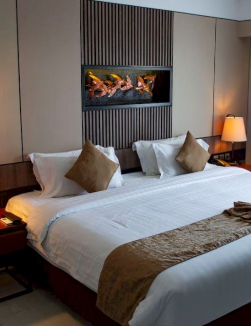 A modern hotel room with a king-size bed, two bedside lamps, decorative cushions, and a small wall-mounted fireplace above the bed.