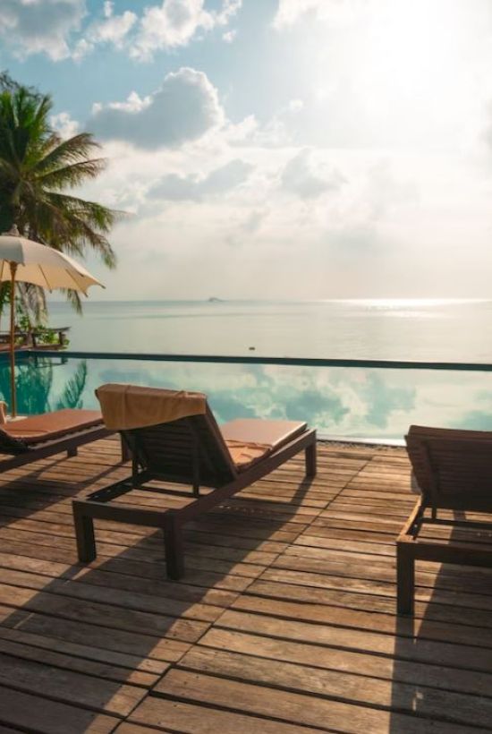A serene poolside scene with lounge chairs, umbrellas, palm trees, and a view of the ocean under a partly cloudy sky.