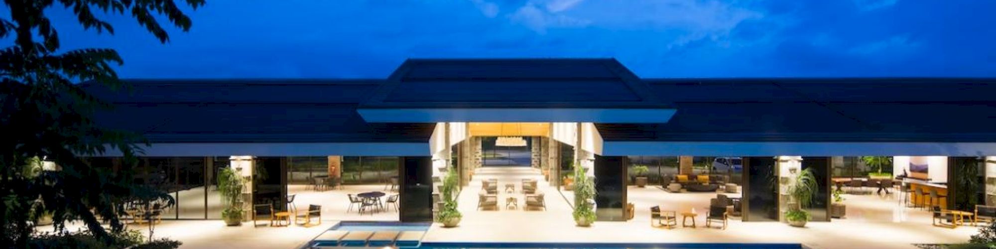 A luxurious modern building with a large, illuminated swimming pool in front, set against a vibrant evening sky with scattered clouds.