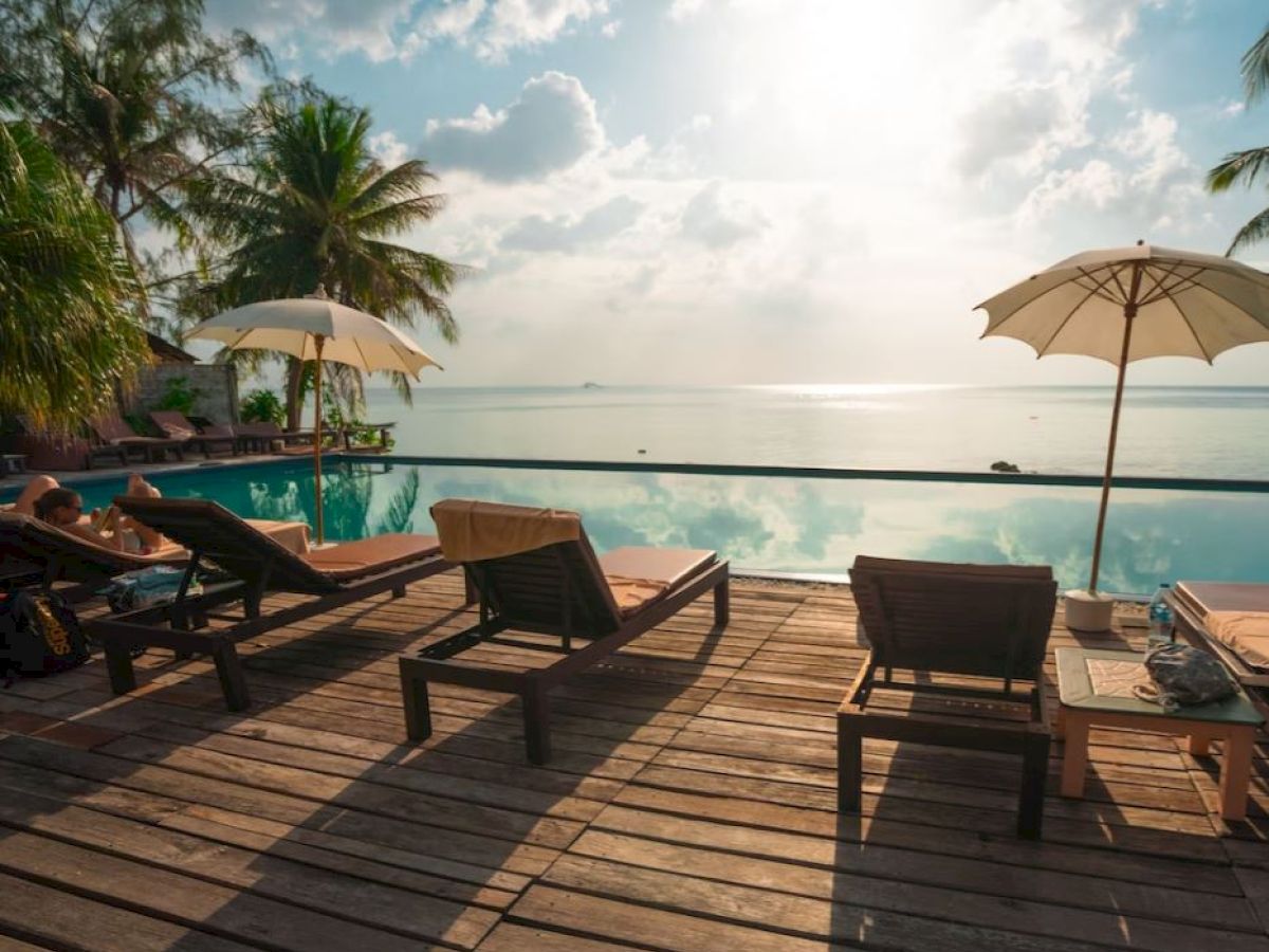 A scenic view of a poolside area with lounge chairs, umbrellas, and palm trees overlooking the ocean under a bright sky.