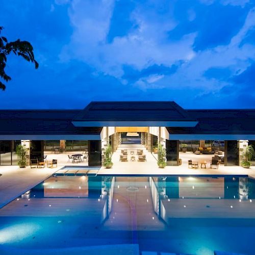 Luxurious outdoor pool area with modern architecture, illuminated at night under a deep blue sky, surrounded by seating and greenery.