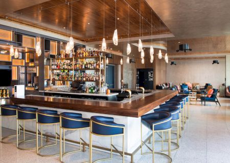 Modern bar with a stocked liquor shelf, pendant lighting, blue bar stools, and seating area, featuring sleek interior design elements.