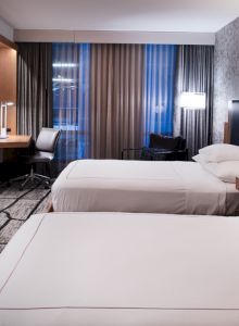 The image shows a modern hotel room with two beds, a flat-screen TV, a work desk, lamps, and patterned wallpaper and carpet.