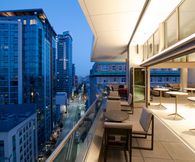 A rooftop terrace with seating overlooks a cityscape of modern buildings at dusk, with streetlights illuminating the scene below.