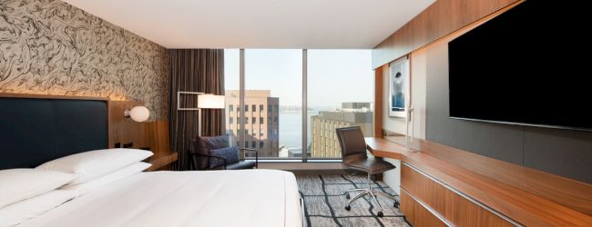 The image shows a modern hotel room with a bed, desk, chair, TV, large window, and city view. The decor is minimal and stylish.