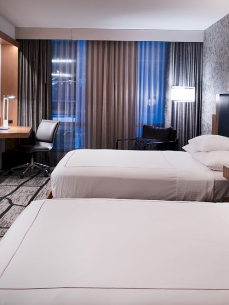 A modern hotel room with two beds, a TV, a desk, and large windows. The decor is sleek with a patterned wall and contemporary lighting.