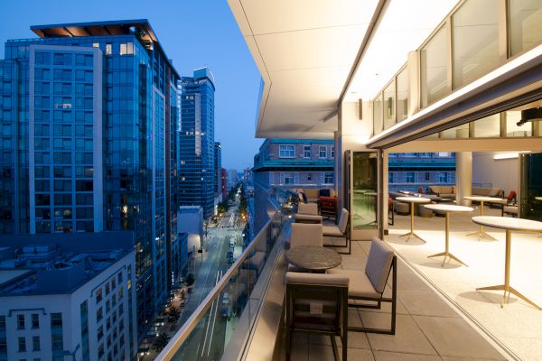 A modern balcony with seating overlooks an urban skyline of tall buildings at dusk, with city lights illuminating the street below.