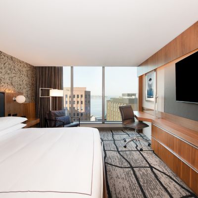 A modern hotel room with a large bed, desk, chair, flat-screen TV, and a window view of buildings outside.