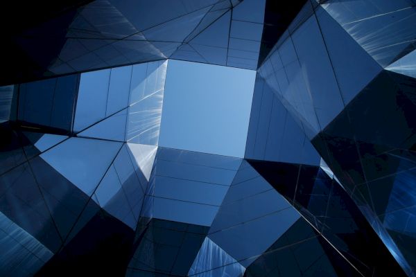 Geometric architecture with reflective blue glass panels creates a dynamic, sky-framed view upward.