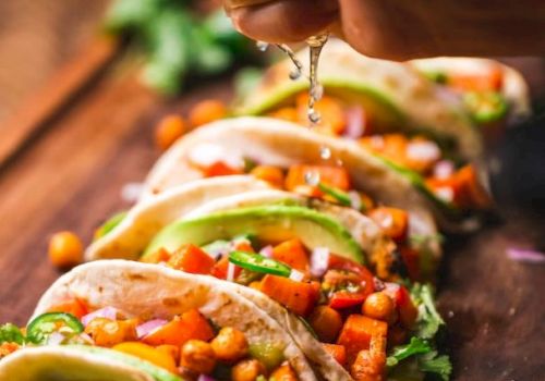 Tacos filled with chickpeas, avocado, and veggies are lined up. A hand is squeezing lime juice over them on a wooden surface.