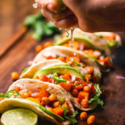 Tacos filled with chickpeas, avocado, and veggies are lined up. A hand is squeezing lime juice over them on a wooden surface.