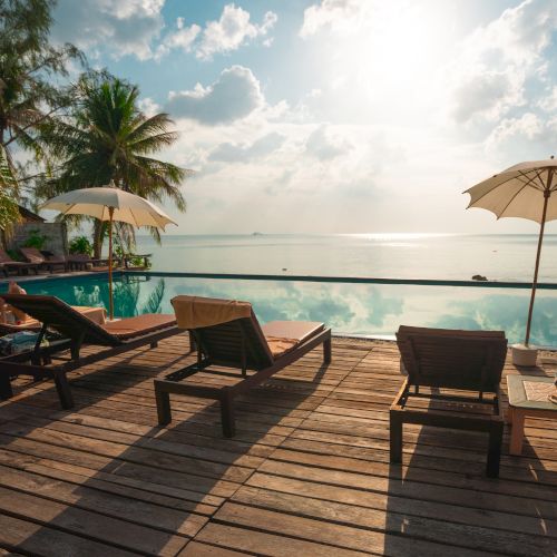 Lounge chairs and umbrellas on a wooden deck overlooking a pool and ocean, with palm trees and a bright sky in the background.