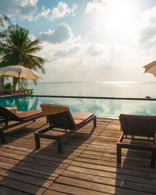 Lounge chairs and umbrellas on a wooden deck overlooking a pool and ocean, with palm trees and a bright sky in the background.
