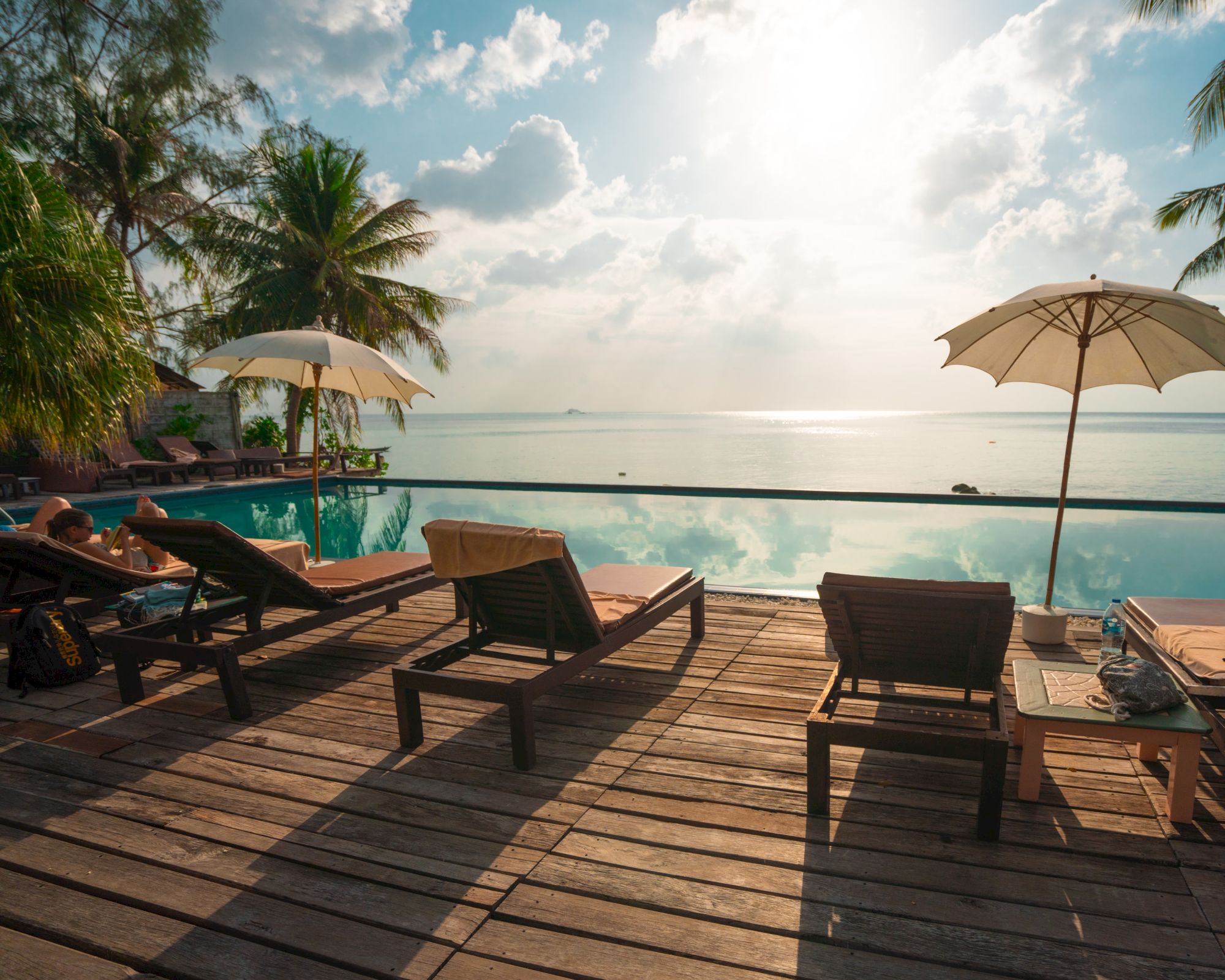 The image shows a tropical beach scene with lounge chairs, umbrellas, and an infinity pool overlooking the ocean under a bright sky.