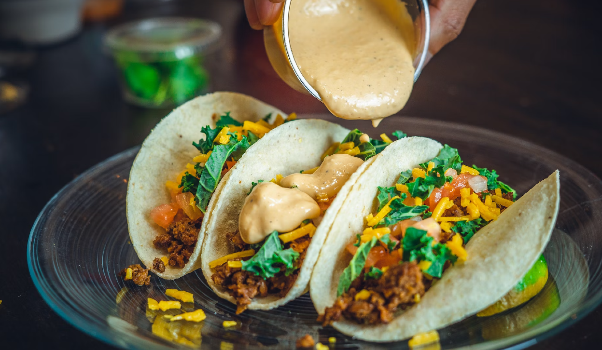 Three tacos filled with ground meat, cheese, lettuce, and tomato are being topped with a creamy sauce on a glass plate.