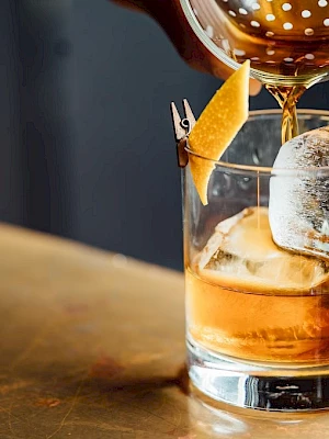 A cocktail being poured into a glass with ice and a lemon twist on a wooden surface, creating a classic and elegant beverage presentation.