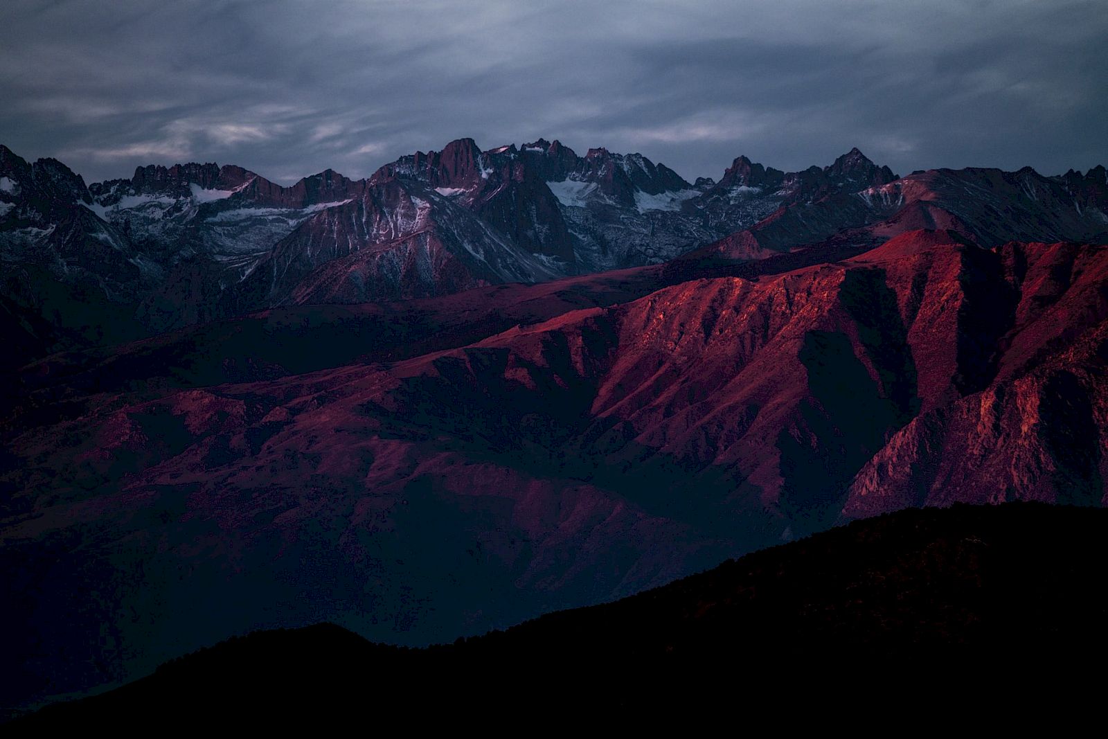 A mountain range bathed in deep red light at sunset, with rugged peaks under a cloudy sky.