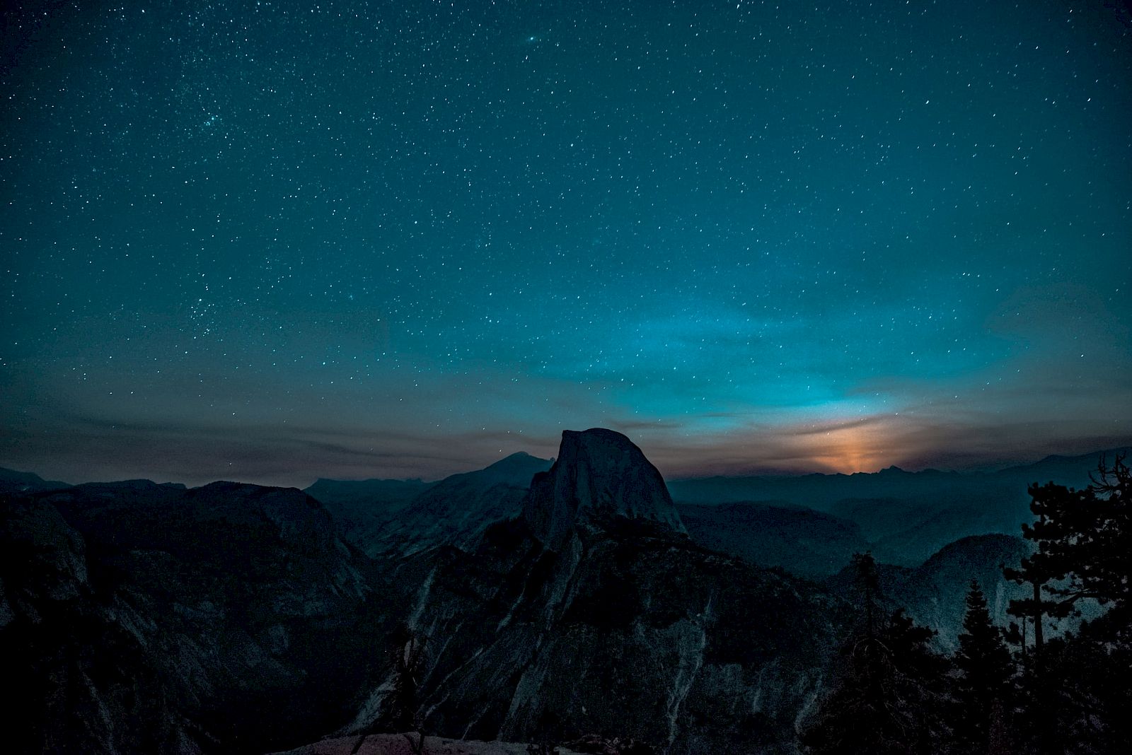 A serene night sky filled with stars over a mountain landscape, with a faint glow, possibly from the moon or sunset, at the horizon.