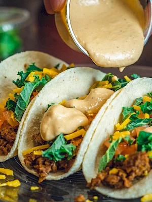 Three tacos with ground beef, lettuce, cheese, tomato, and sauce being poured over them on a plate.