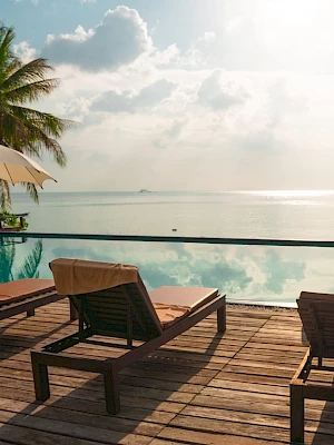 Poolside view with lounge chairs, umbrellas, and palm trees overlooking a serene ocean under a bright sky, creating a relaxing atmosphere.