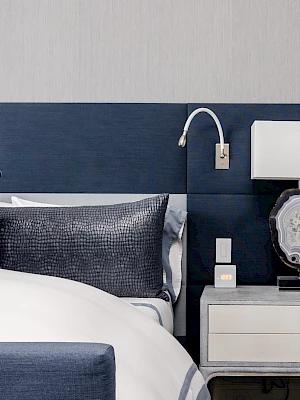 A modern bedroom with a blue padded headboard, white bedding, a nightstand, lamp, and wall-mounted reading light.