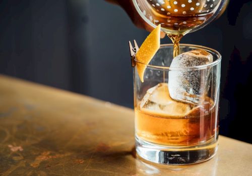 A person is pouring a brown cocktail into a glass with ice and a lemon twist, using a strainer, on a wooden surface.