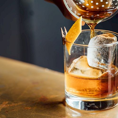 A person is pouring a brown cocktail into a glass with ice and a lemon twist, using a strainer, on a wooden surface.