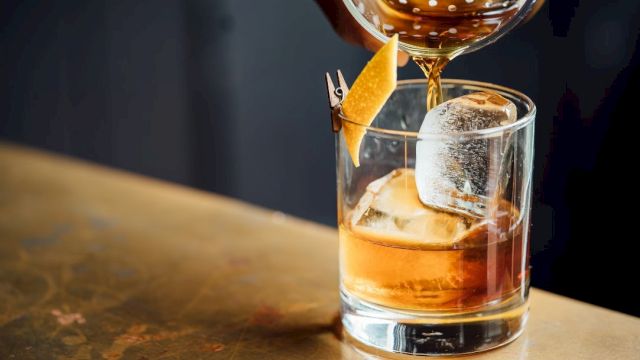 A person is pouring a brown cocktail into a glass with ice and a lemon twist, using a strainer, on a wooden surface.