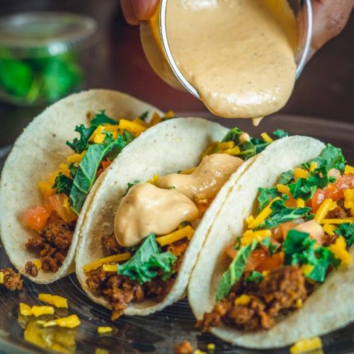 Three tacos with meat, shredded cheese, lettuce, and a sauce being poured over them on a plate.
