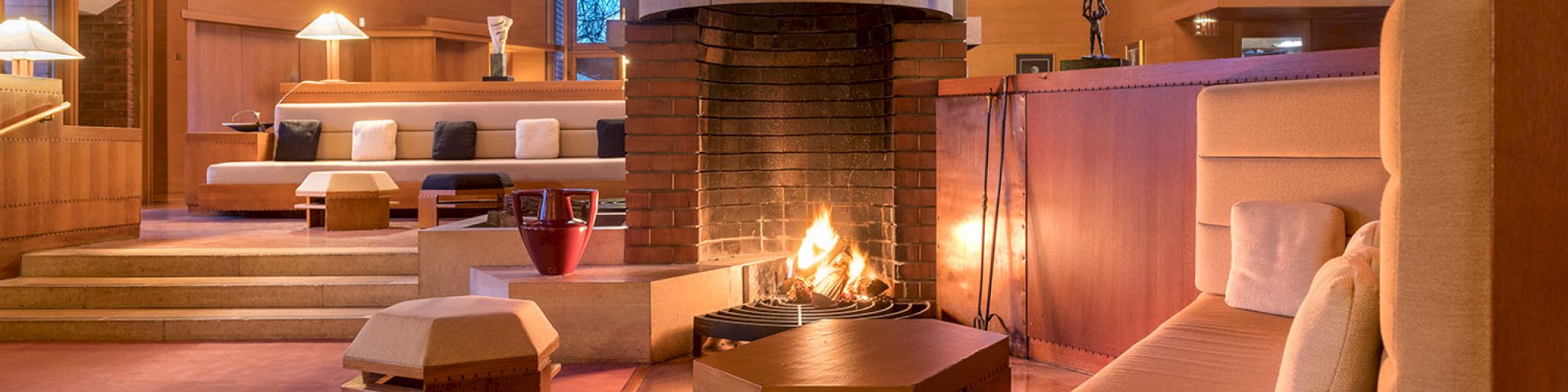 The image shows a cozy living room with wooden interiors, a central fireplace, and cushioned seating, creating a warm, inviting atmosphere.