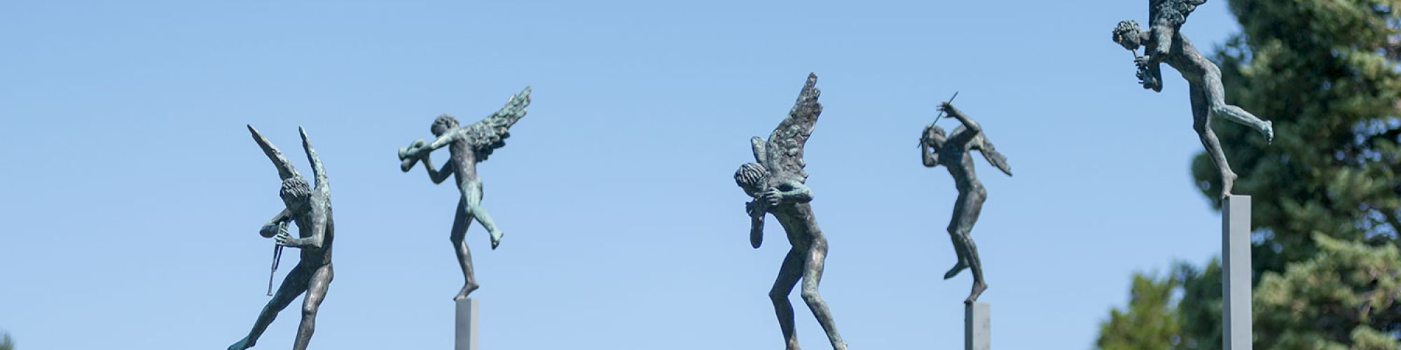 The image features sculptures of winged figures playing instruments, positioned on tall poles against a backdrop of trees and a building.