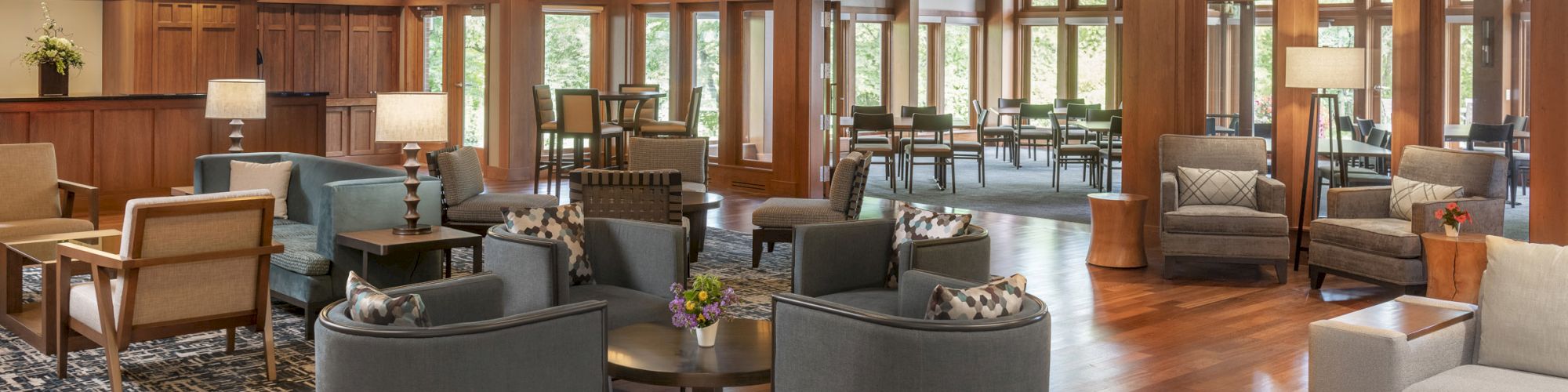 A spacious, warmly-lit hotel lobby with wooden floors, modern seating areas, and large windows. Numerous lights hang from the ceiling.
