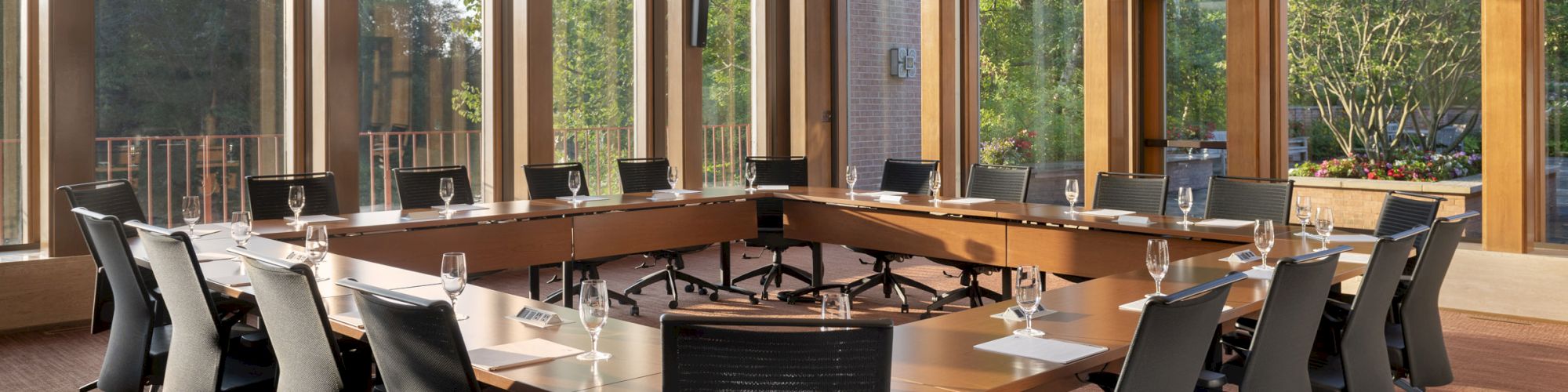 The image shows a conference room with a large U-shaped table, black chairs, large windows, and a view of greenery outside.