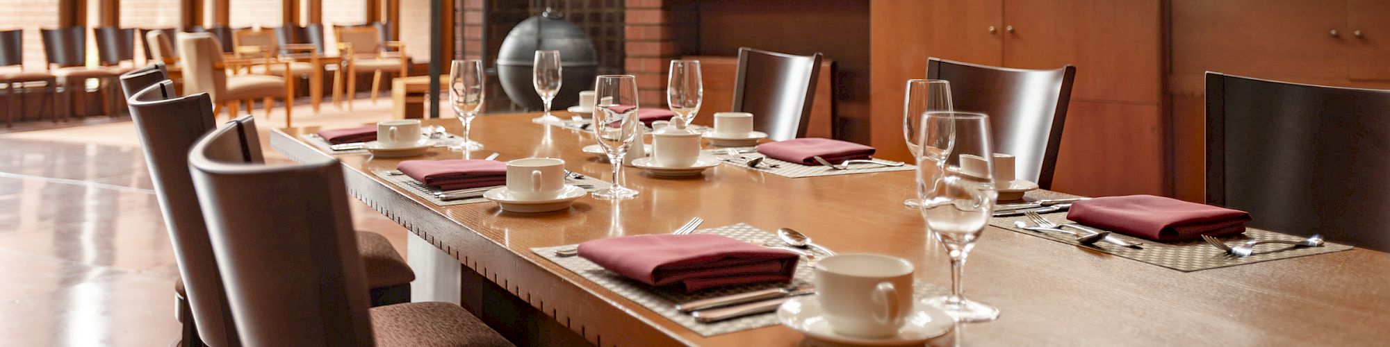 A modern dining room with a wooden table set with glasses and napkins, featuring brick walls and large windows.