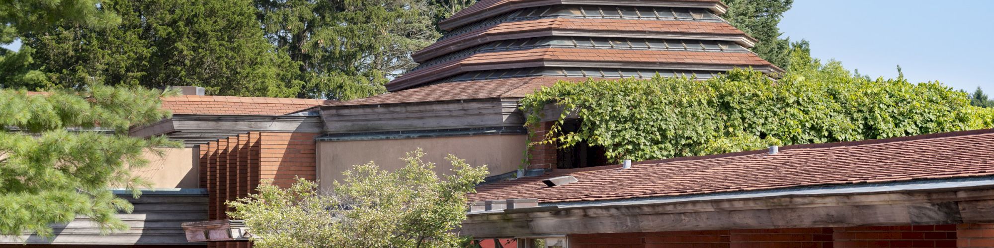 The image shows a building with a unique, layered roof structure, surrounded by trees and plants, with planters along a red walkway.