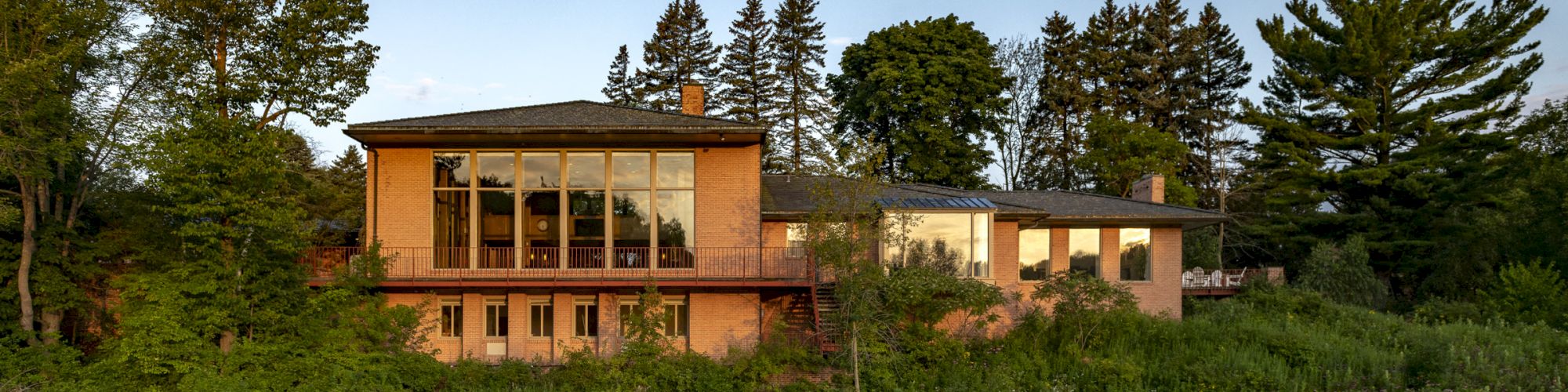 A modern house with large windows is situated beside a calm river, surrounded by lush greenery and reflections in the water.