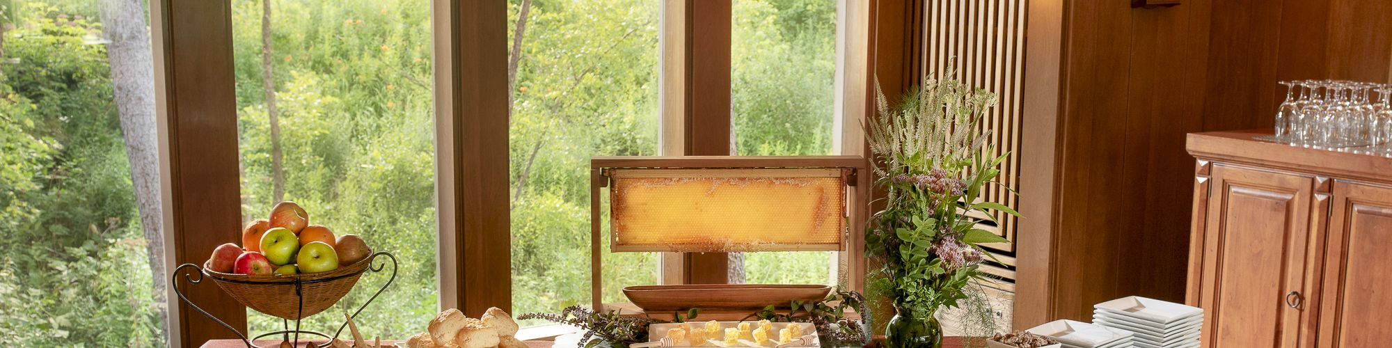 A buffet table with various foods, fruits, and utensils is set up indoors beside large windows with a scenic view.