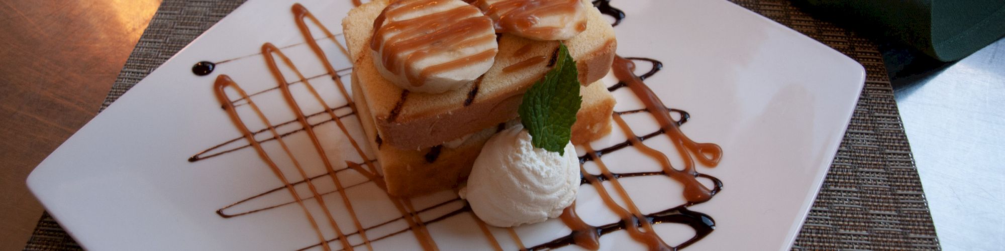 A dessert on a square plate with caramel and chocolate drizzle, topped with whipped cream and mint, rests on a woven placemat.