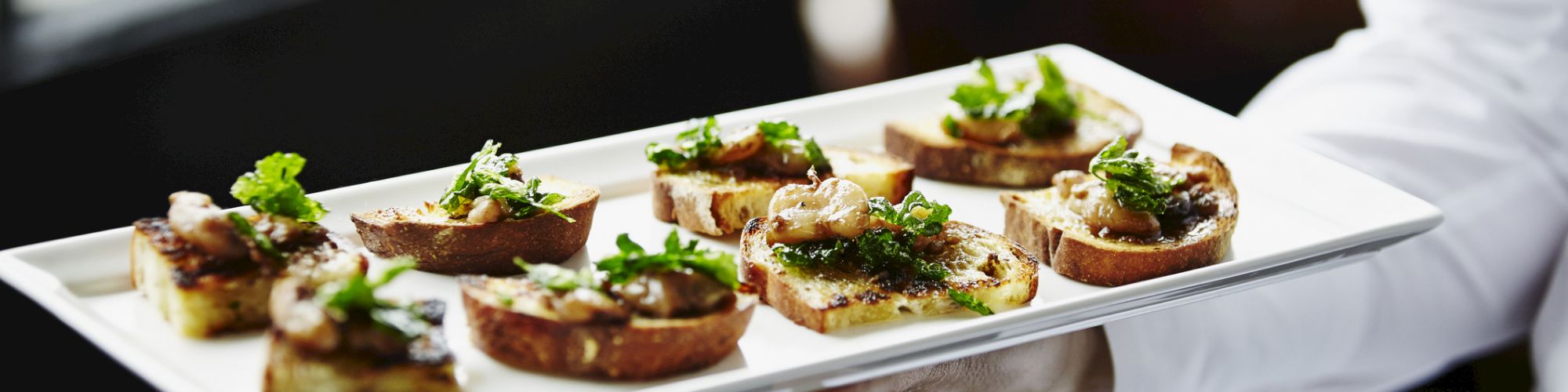 A person holds a tray with elegant bruschetta topped with herbs and garnishes, ready to be served in a light-filled setting.