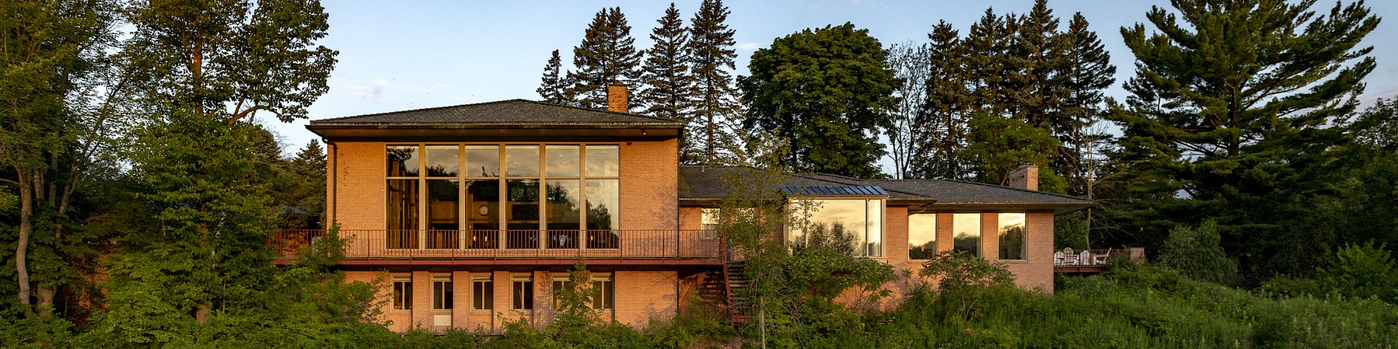 A modern house with large windows is reflected in a calm river, surrounded by lush greenery and trees under a partly cloudy sky.