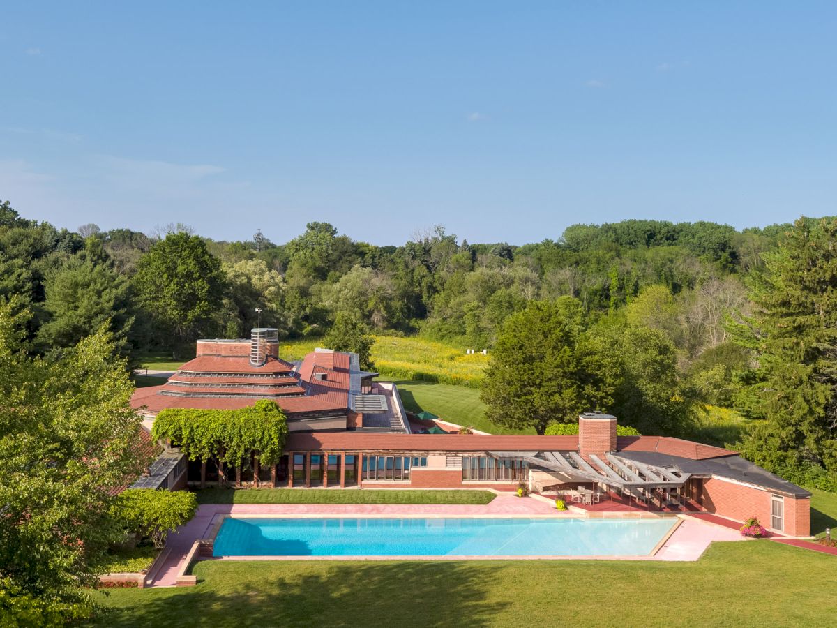 A large house with a unique design, surrounded by trees and greenery, featuring an outdoor swimming pool under a clear blue sky.