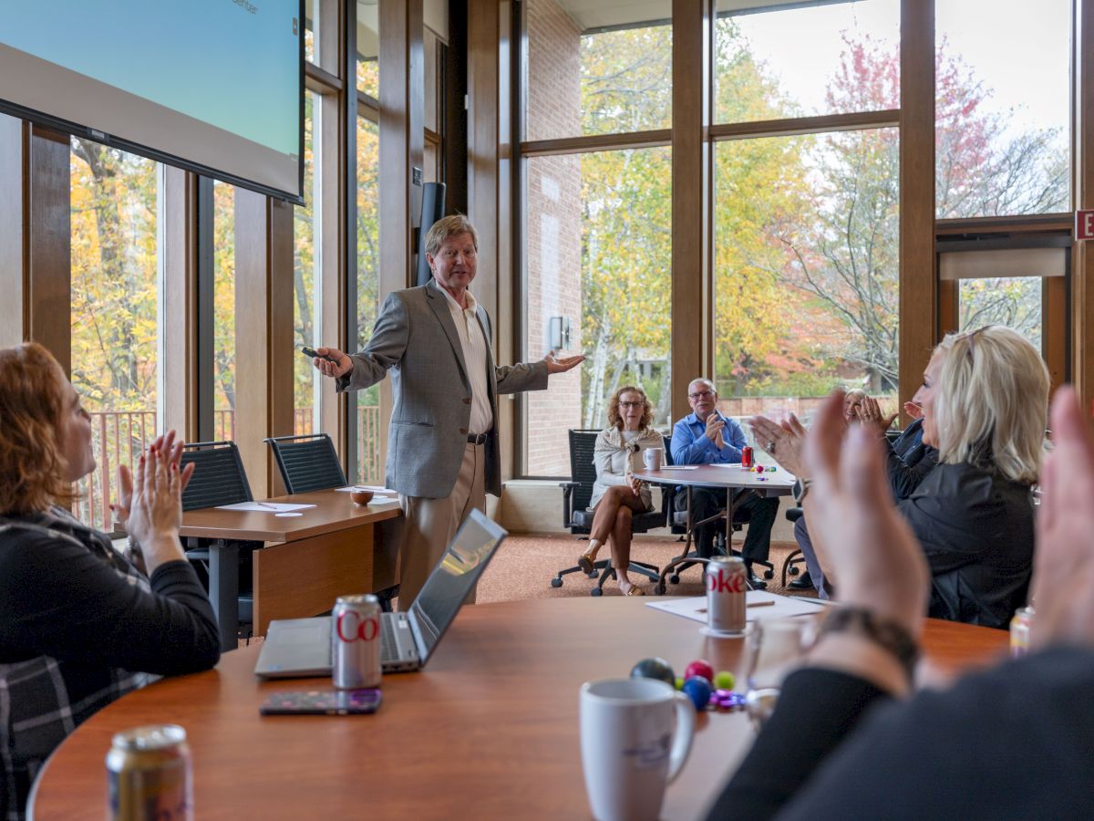 A man in a suit presents in a bright conference room while colleagues cheer and take notes, a collaborative meeting in progress.