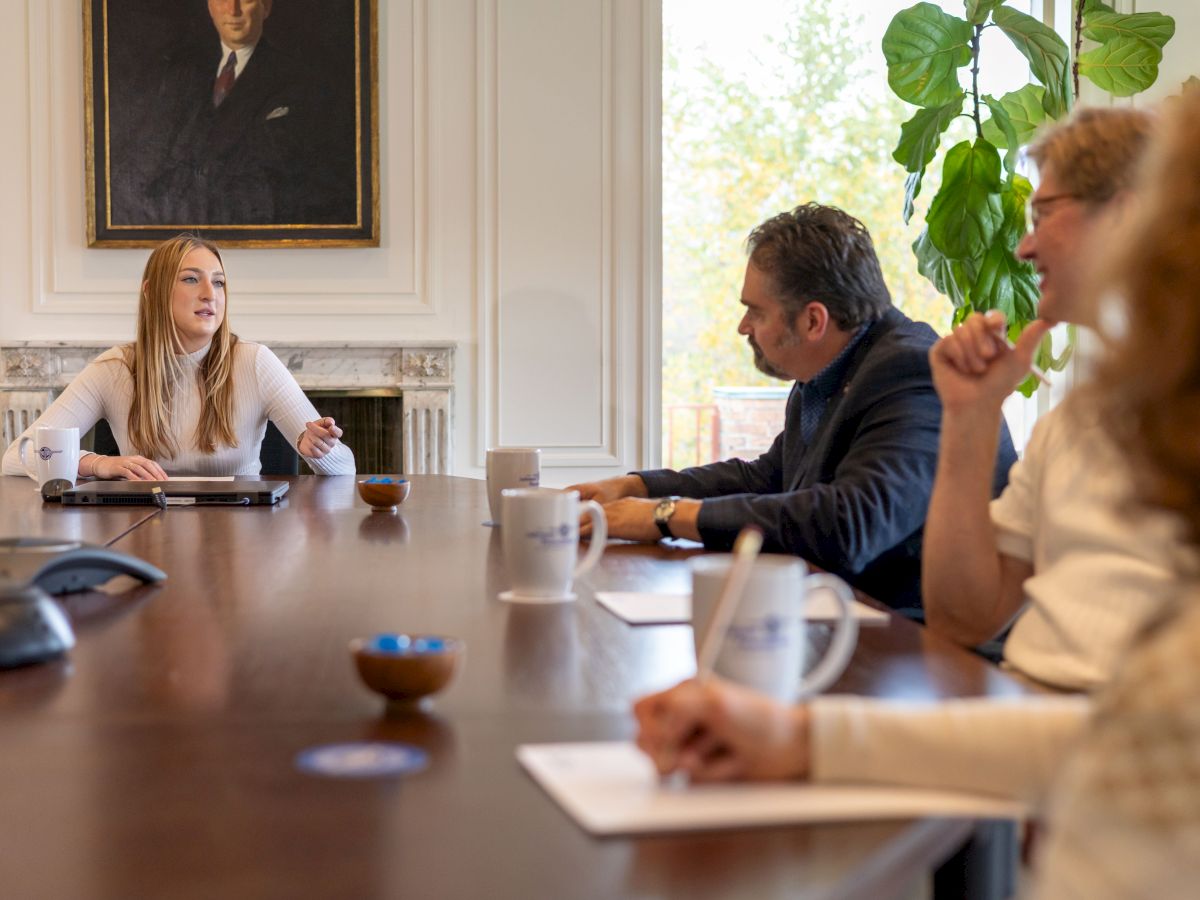 A group of professionals sits around a conference table in a bright office, discussing with notebooks and mugs in view, a plant and portrait in the background. End.