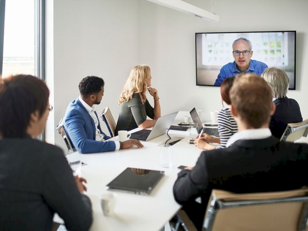 People in a meeting room are watching a person on a screen, likely participating in a video conference.