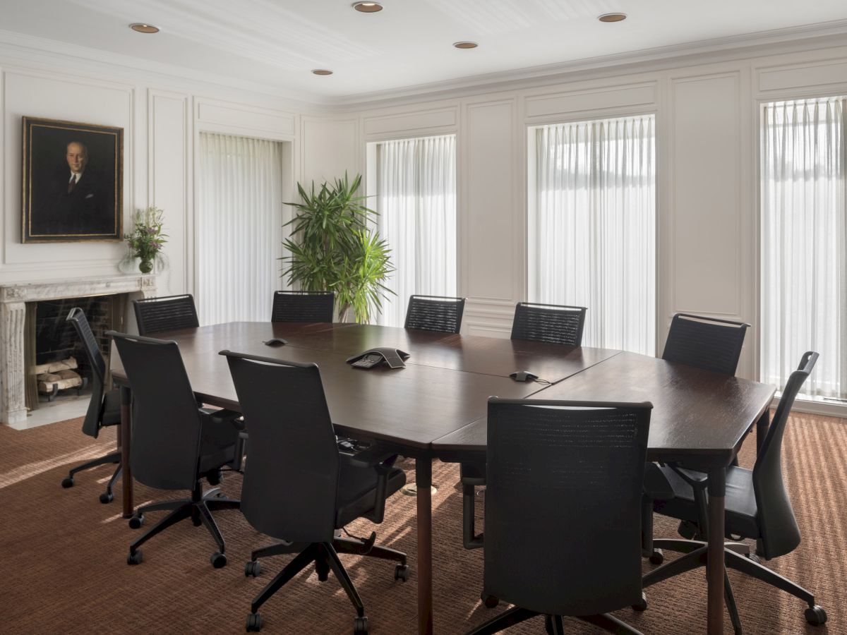 The image shows a conference room with a large oval table, black chairs, a plant, and a portrait on the wall.