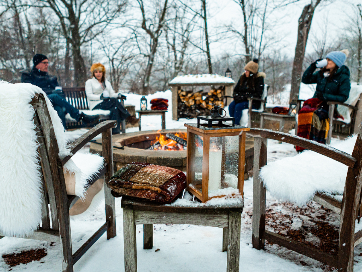 Four people sit outdoors around a fire pit on a snowy day, with wooden benches and cozy throws. Snow covers the ground and trees.
