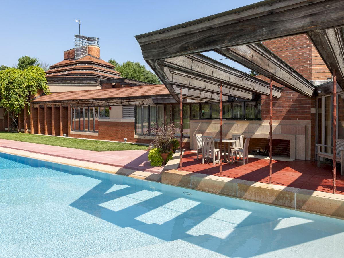 A modern house with a swimming pool, featuring angled roofs and large windows, surrounded by trees and blue sky.